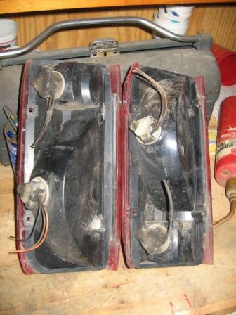 1993 Ford Bronco - A pair of removed rear taillight housings laid face-down on a wooden workbench, showing the dusty black plastic reflector cavities, socket…