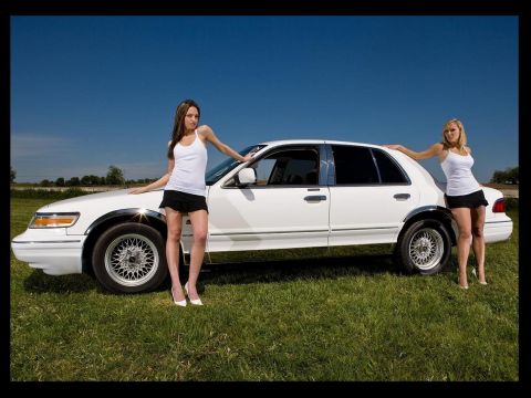 1997 Mercury Grand Marquis - A white 1997 Mercury Grand Marquis is shown with two women posing beside it on a grassy field under a clear blue sky.