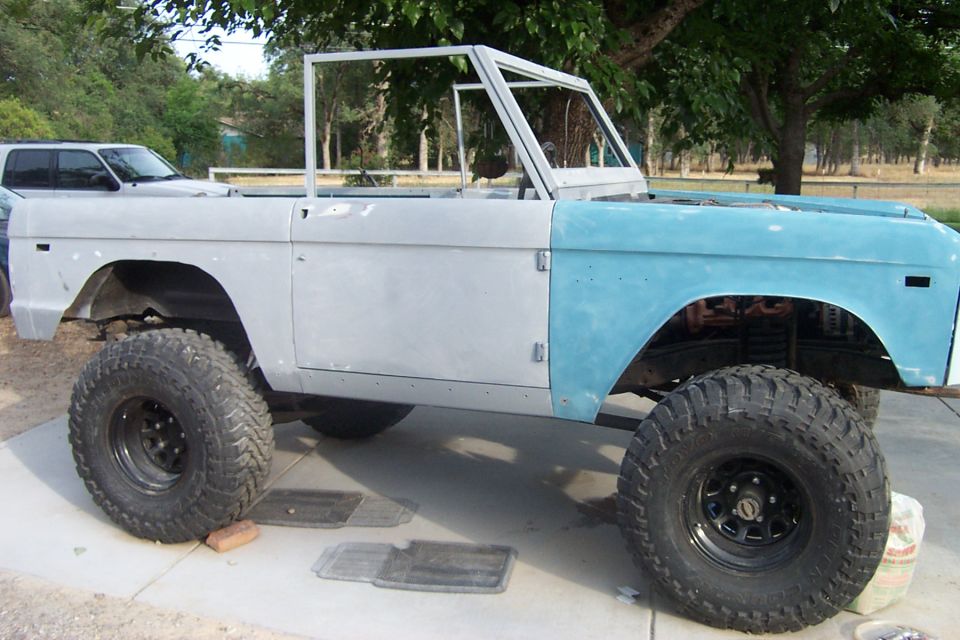 Unfinished 1976 Ford Bronco body, primer gray and light blue, on large tires, side view