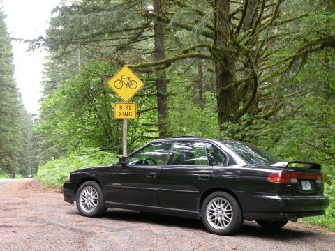 1998 Subaru Legacy - A black 1998 Subaru Legacy sedan is parked on a dirt road in a forest, with a bike crossing sign visible.
