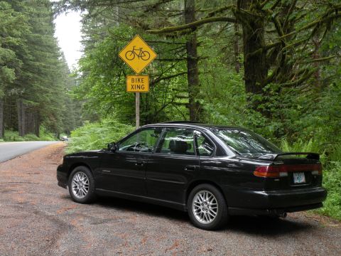 1998 Subaru Legacy - A black 1998 Subaru Legacy sedan is parked on a gravel road in a forest area.