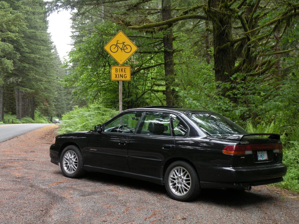Black 1998 Subaru Legacy sedan parked on a gravel shoulder next to a forest road with a bike crossing sign