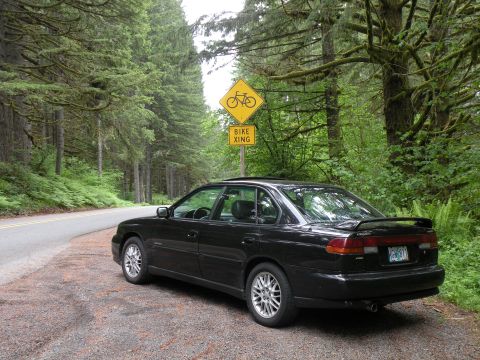 1998 Subaru Legacy - A black sedan is parked on the side of a road in a wooded area, with a bike crossing sign visible.
