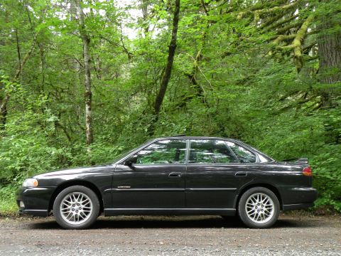 1998 Subaru Legacy - A black 1998 Subaru Legacy sedan is parked on a gravel road surrounded by lush green trees.
