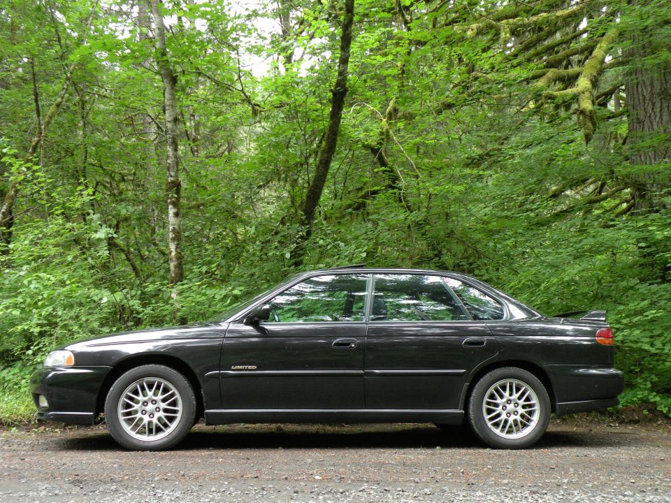 Black 1998 Subaru Legacy sedan, side view, parked on a gravel road with trees in the background