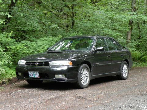 1998 Subaru Legacy - This black 1998 Subaru Legacy sedan is parked on a dirt road surrounded by trees.