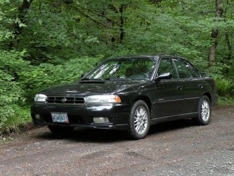 1998 Subaru Legacy - Black 1998 Subaru Legacy sedan parked on a gravel road surrounded by trees.