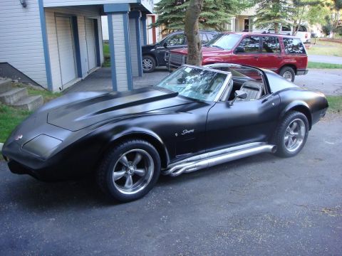 1975 Chevrolet Corvette - A matte black 1975 Chevrolet Corvette Stingray is parked in a driveway next to other vehicles.