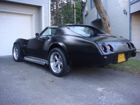 1975 Chevrolet Corvette - Matte black 1975 Chevrolet Corvette with custom wheels and exhaust pipes parked in front of a garage.