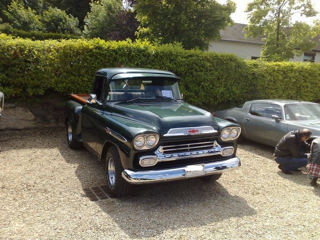 Green 1958 Chevrolet pickup truck with chrome grille and headlights, parked on gravel next to a silver car