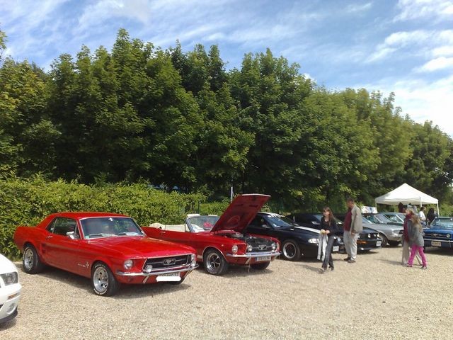 Red 1967 Ford Mustang coupe parked next to a red convertible with hood open; other cars and people in background