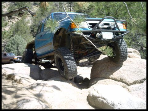 1993 Ford Bronco - A heavily modified blue pickup truck navigates a rocky trail, with another vehicle visible in the background.