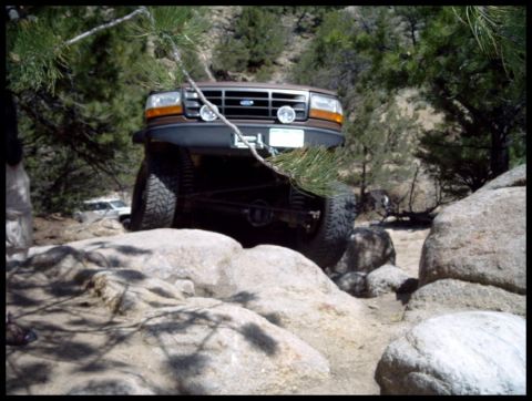 1993 Ford Bronco - A brown 1993 Ford Bronco is shown navigating a rocky trail, with another vehicle visible in the background.