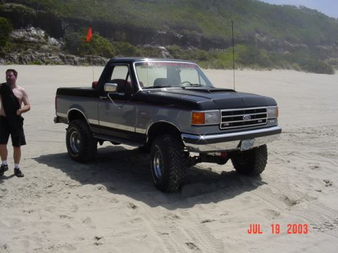1989 Ford Bronco - A two-tone Ford Bronco with off-road tires sits on a sandy beach, with a man and a red flag visible.