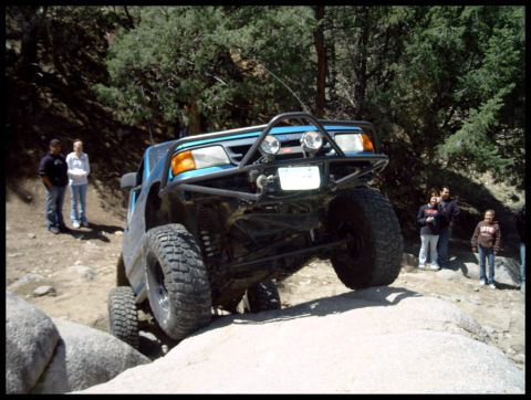 1993 Ford Bronco - A blue off-road SUV is shown traversing a challenging rocky path with onlookers present.