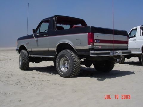 1989 Ford Bronco - A lifted Ford Bronco sits on a sandy beach next to another white SUV under a clear blue sky.