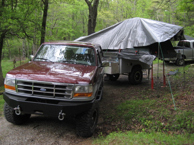 Lifted maroon OBS Ford truck with black tube bumper at wooded campsite beside tarp-covered trailer