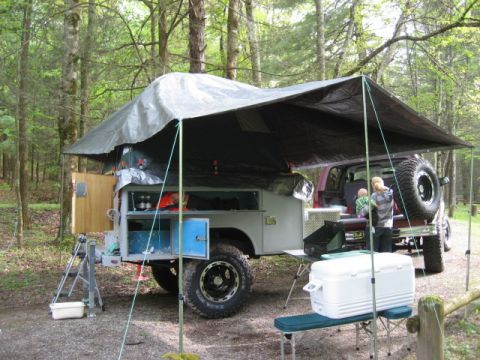 1996 Ford Bronco - A camper trailer is set up in a wooded area with a vehicle attached, showing a spare tire mounted on the rear.