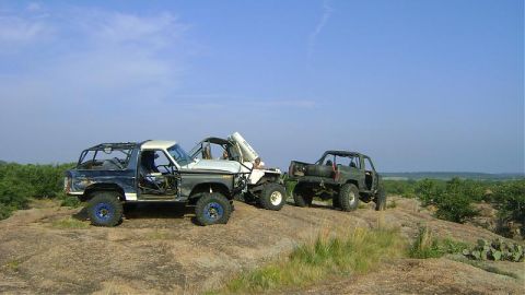 1986 Ford Bronco - Modified off-road vehicles are parked on a rocky terrain with sparse vegetation.
