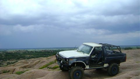 1986 Ford Bronco - This 1986 Ford Bronco, modified for off-roading with a roll cage, sits on a rocky outcrop under a cloudy sky.