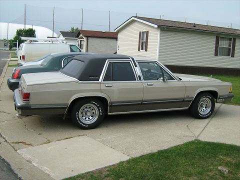 1988 Mercury Grand Marquis - Tan 1980s-90s Mercury Grand Marquis four-door with padded vinyl roof and chrome trim, parked in profile in a residential driveway by a beig…