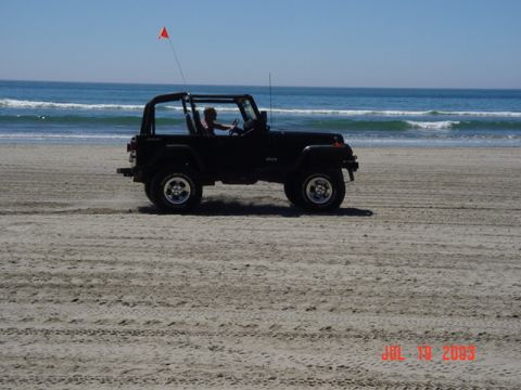 1989 Ford Bronco - A black Jeep Wrangler drives along a sandy beach on a sunny day, with the ocean waves in the distance.