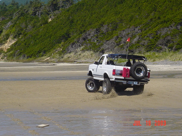 Lifted white Ford Bronco with bikini top and rear spare carrier driving on a beach