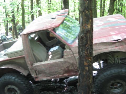 1991 Ford Bronco - A damaged red 1991 Ford Bronco is stuck in a wooded area with other vehicles visible in the background.
