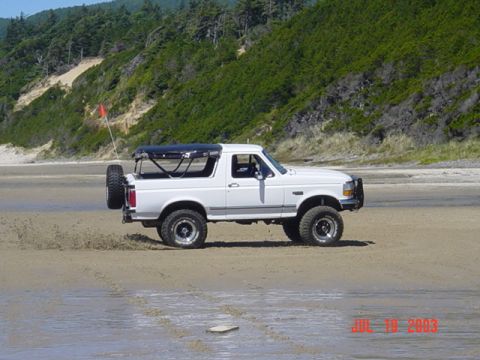 1989 Ford Bronco - White Ford Bronco with a soft top and spare tire on the back driving on a beach.