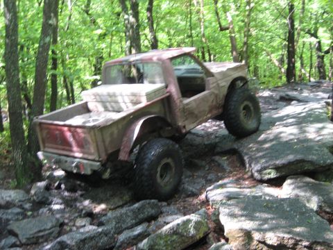 1991 Ford Bronco - An off-road vehicle with large tires is shown navigating a challenging rocky trail through a wooded area.