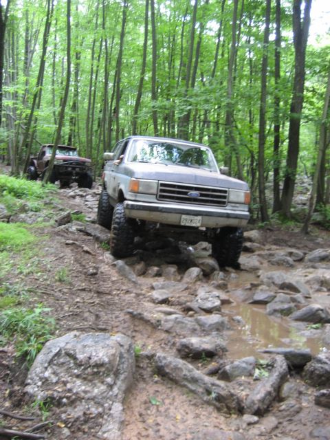 1991 Ford Bronco - Two off-road vehicles navigate a rocky, muddy path through a dense forest during an event.