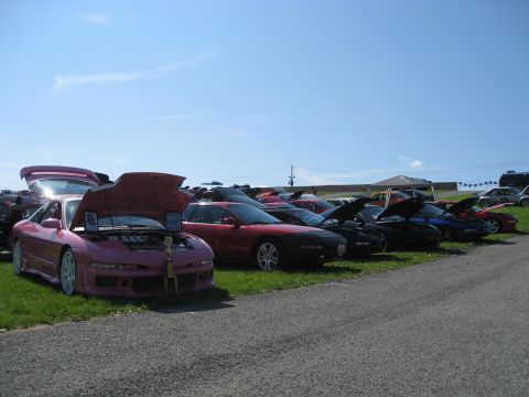 1994 Ford Probe - A line of cars are parked on a grassy area with their hoods open under a clear blue sky.
