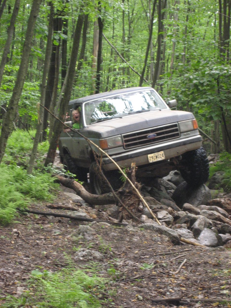 Ford Bronco navigating a rocky trail in the woods. Driver is visible and giving a peace sign