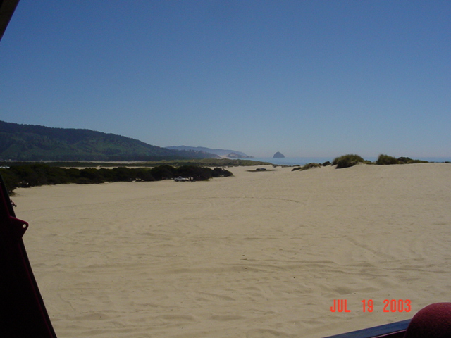 Sandy dunes with tire tracks near the coast, haystack rock in the distance