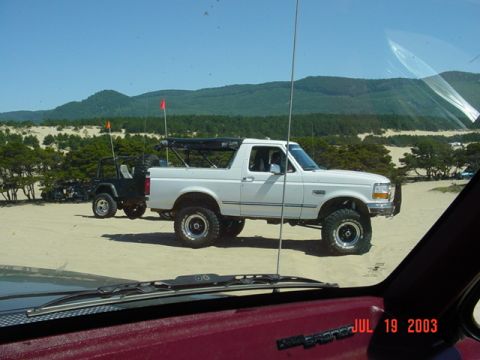 1989 Ford Bronco - A white Ford Bronco and a black Jeep are parked on sand dunes with trees and hills in the background.
