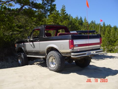 1989 Ford Bronco - This lifted Ford Bronco is parked on a sandy terrain with trees and a clear blue sky overhead.