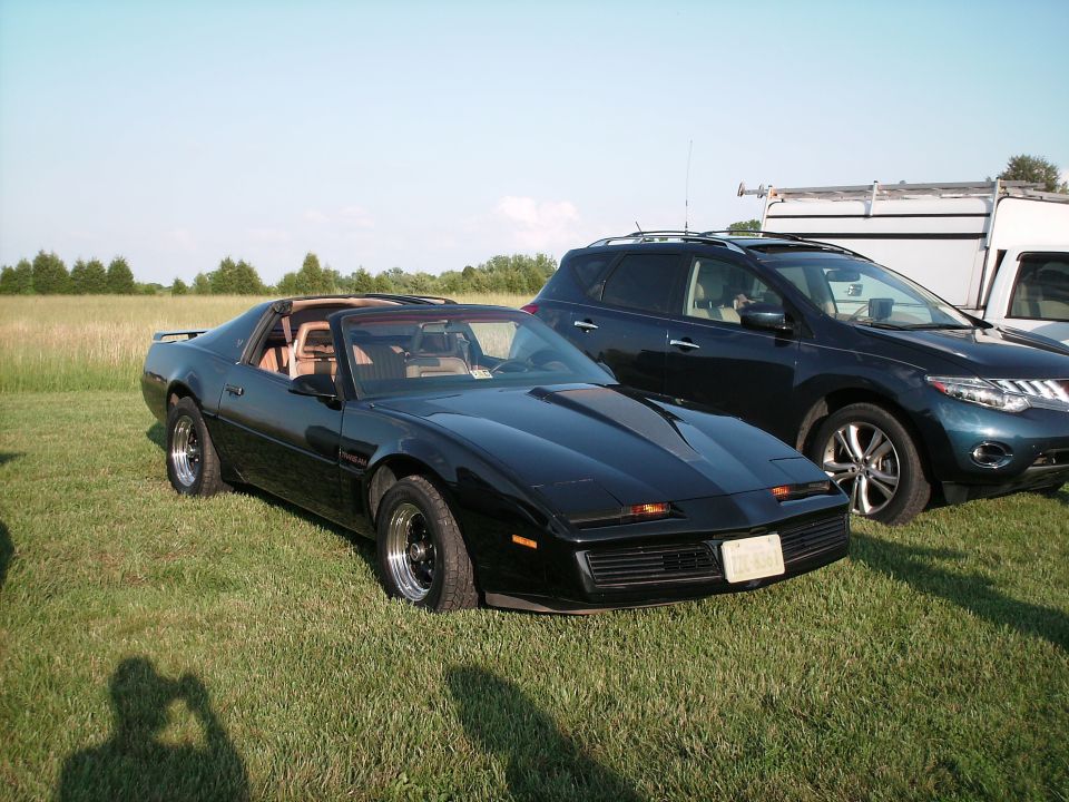 Black Pontiac Firebird parked on grass next to a dark SUV and a white camper