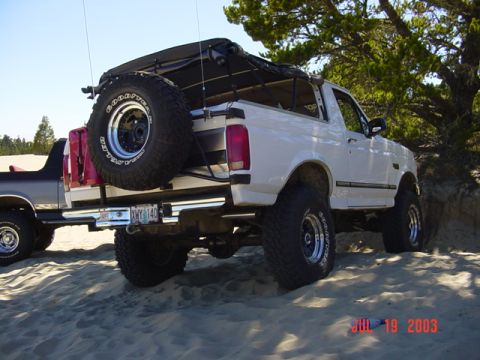 1989 Ford Bronco - A white Ford Bronco with a soft top and spare tire is parked on a sandy dune next to another vehicle.