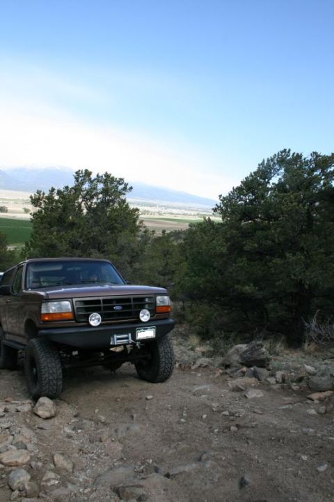 1993 Ford Bronco - A brown 1993 Ford Bronco navigates a , rocky terrain with trees and mountains in the background.