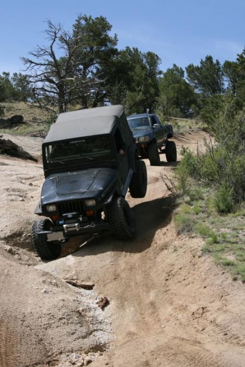 1993 Ford Bronco - A black Jeep Wrangler TJ navigates a steep dirt trail, followed by a blue pickup truck, under a clear blue sky.