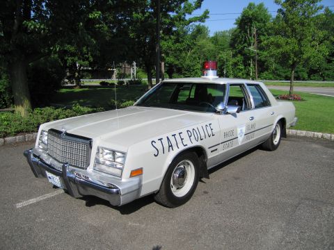 1991 Ford LTD Crown Victoria - cream-colored Rhode Island State Police Chrysler sedan with a single red beacon is parked on a green lawn-edged lot.