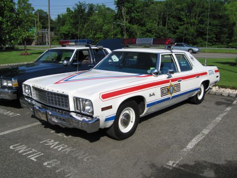 1991 Ford LTD Crown Victoria - white vintage Plymouth sedan with red and blue stripes, marked State Police with a star emblem, is parked in a tree-lined lot.