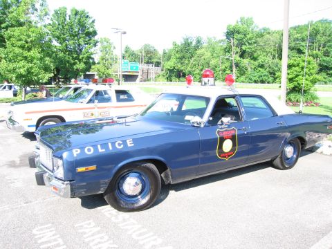 1991 Ford LTD Crown Victoria - dark blue vintage POLICE sedan with a gold badge emblem and black steel wheels sits beside other vintage cruisers in a lot.