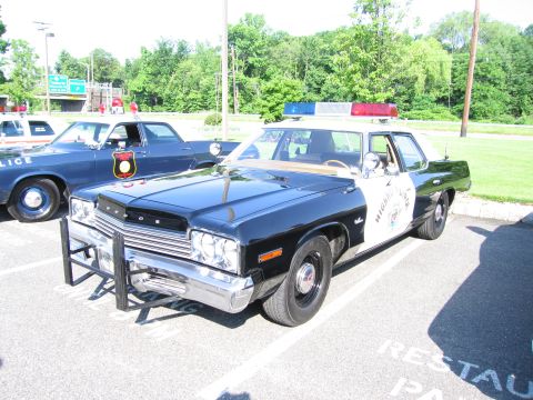 1991 Ford LTD Crown Victoria - black-and-white vintage Dodge Highway Patrol sedan fitted with a push bar is parked beside other classic cruisers on a lot.