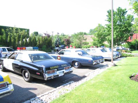 1991 Ford LTD Crown Victoria - lineup of vintage police sedans, including a black Highway Patrol car, parked along a grassy, tree-lined lot.