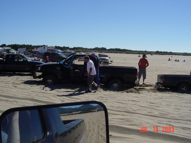 Black pickup buried in beach sand being hooked to a tow strap while bystanders help, dated July 19 2003