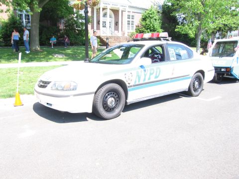1991 Ford LTD Crown Victoria - white four-door police sedan marked NYPD parked on a street in front of a brick library building at a car show.