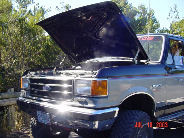 Gray and blue Ford Bronco with hood up and steam rising, parked on a sandy trail among pines