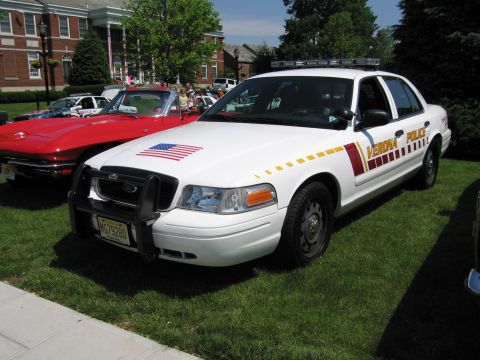 1991 Ford LTD Crown Victoria - white Verona Police sedan with a US flag decal, push bumper and roof lightbar parked on grass at an outdoor car show.