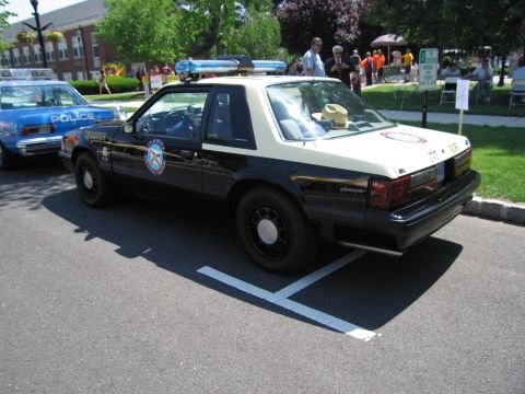 1991 Ford LTD Crown Victoria - black-and-tan Ford Mustang police coupe with circular door emblem and roof lightbar parked at an outdoor car show.